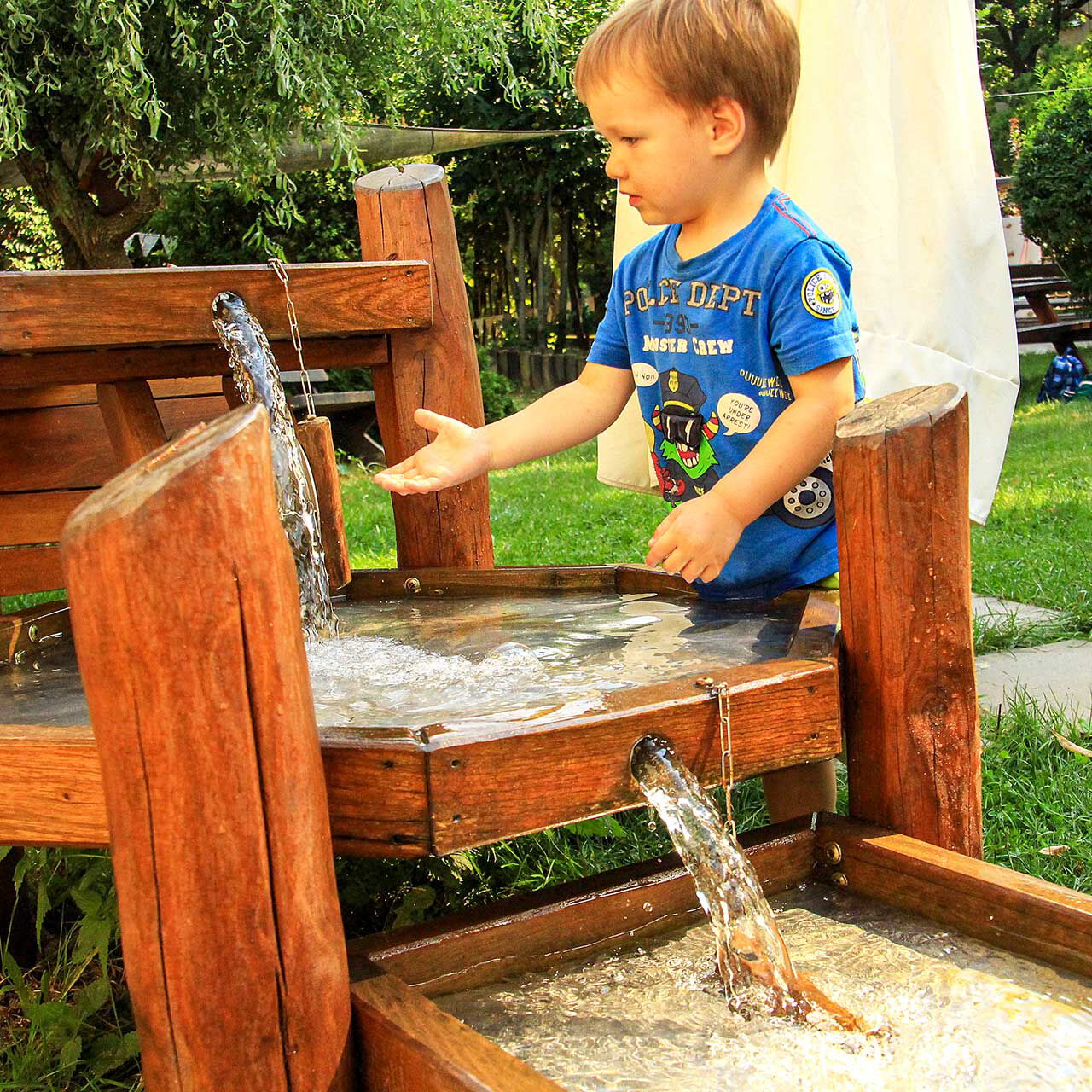 Die Matschanlage mit Wasserspiel aus Holz im Außengelände. Ein Junge spielt mit dem Wasser.