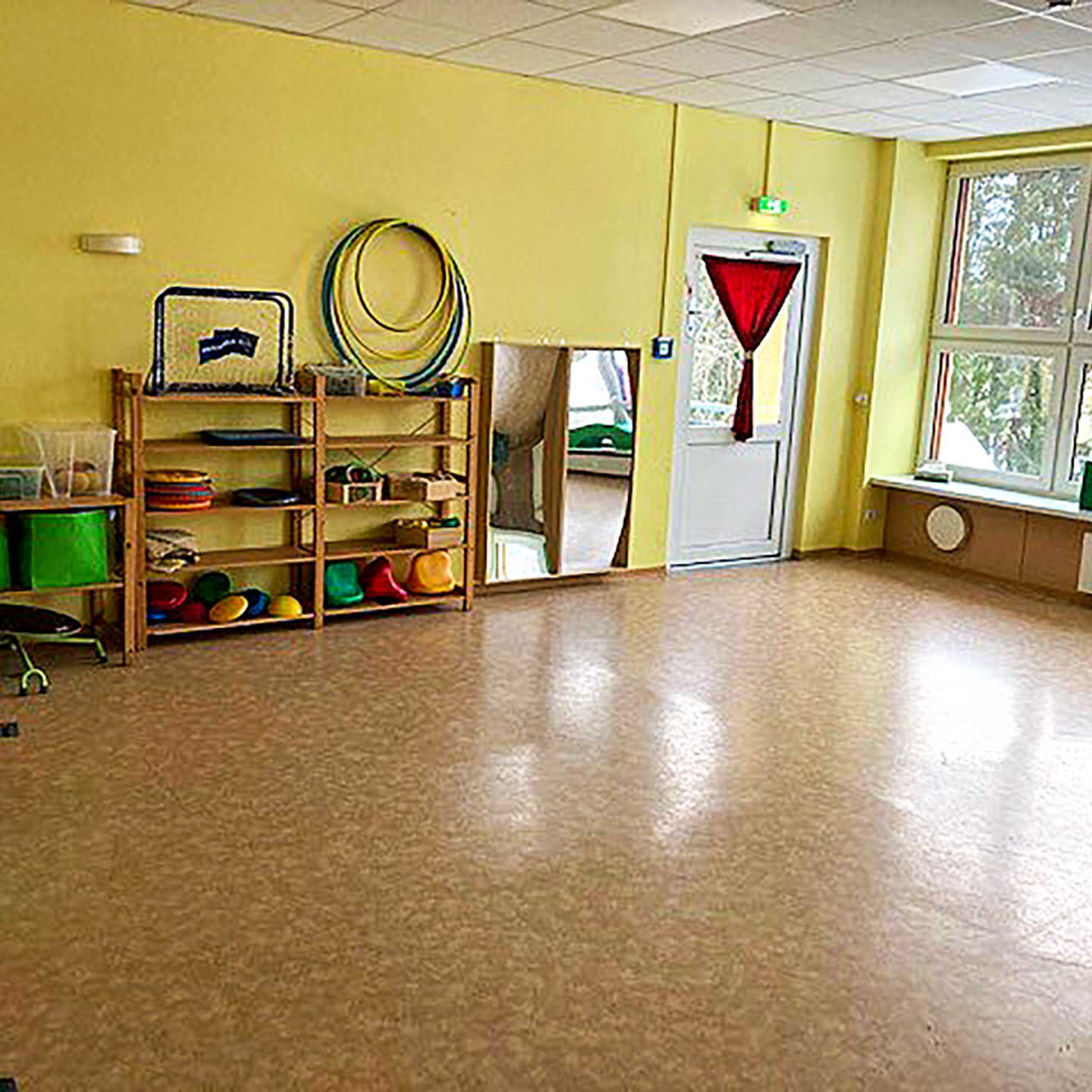 Der Bewegungsraum umfasst einen großen leeren Spielbereich und an den Wänden befinden sich kleine Holzregale mit diversen Sport- und Spielgeräten.