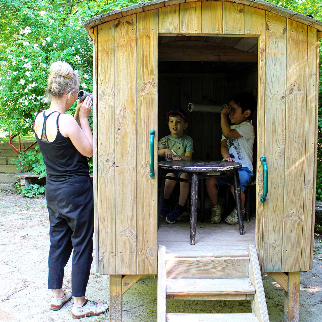 Ein Bauwagen aus Holz im Außengelände. Zwei Kleinkinder sitzen im Inneren und von außen schaut eine Erzieherin durch das Fenster nach innen.