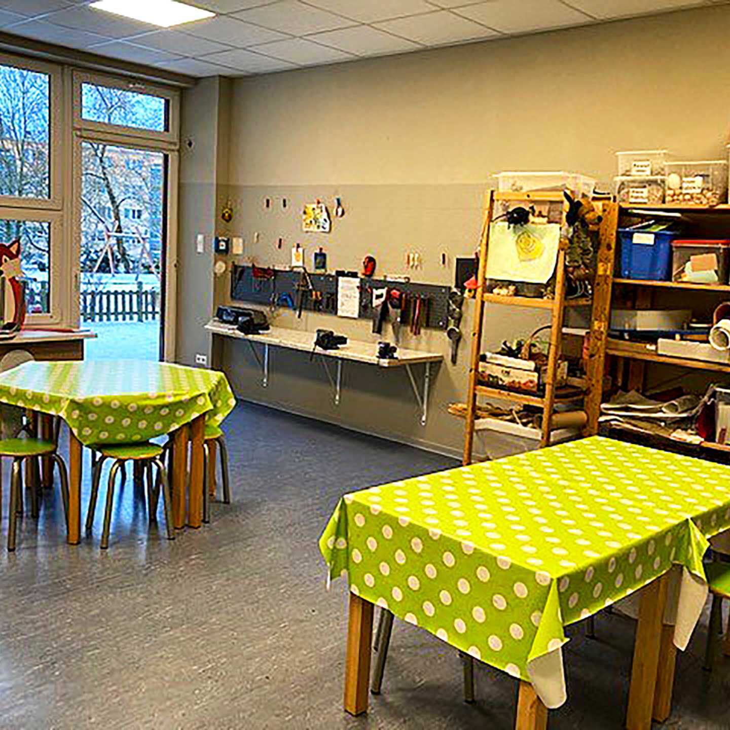 Das Atelier in der Kita besteht aus zwei Tischen mit Stühlen, Holzregalen für Bastelmaterialien und einer Arbeitsplatte an der Wand mit vielen unterschiedlichen Werkzeugen.