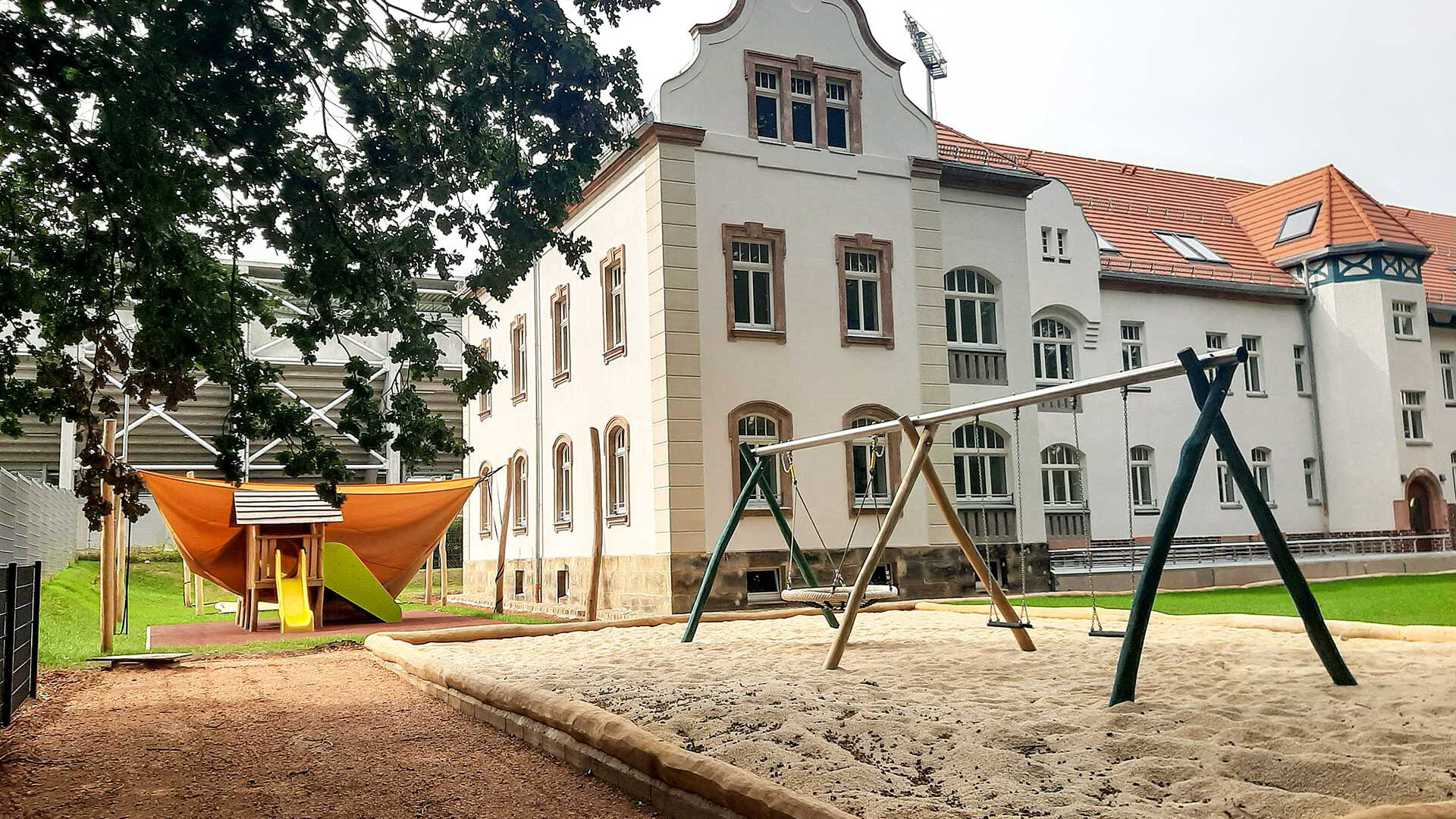 Das Außengelände mit einer Nestschaukel, zwei einzelnen Schaukeln im Vordergrund und das Naturkinderhaus mit einem Spielturm mit Rutsche im Hintergrund.