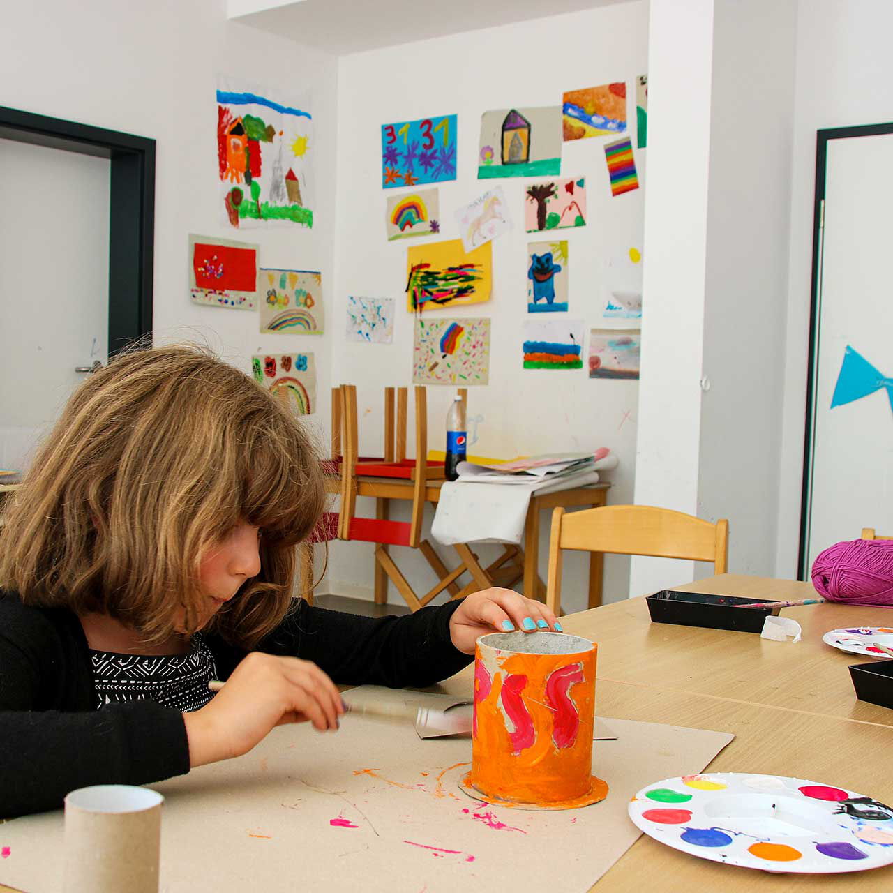 Im Kreativzimmer sitzt ein Mädchen und bemalt eine Dose. Im Hintergrund an der Wand sind viele Kunstwerke zu sehen.