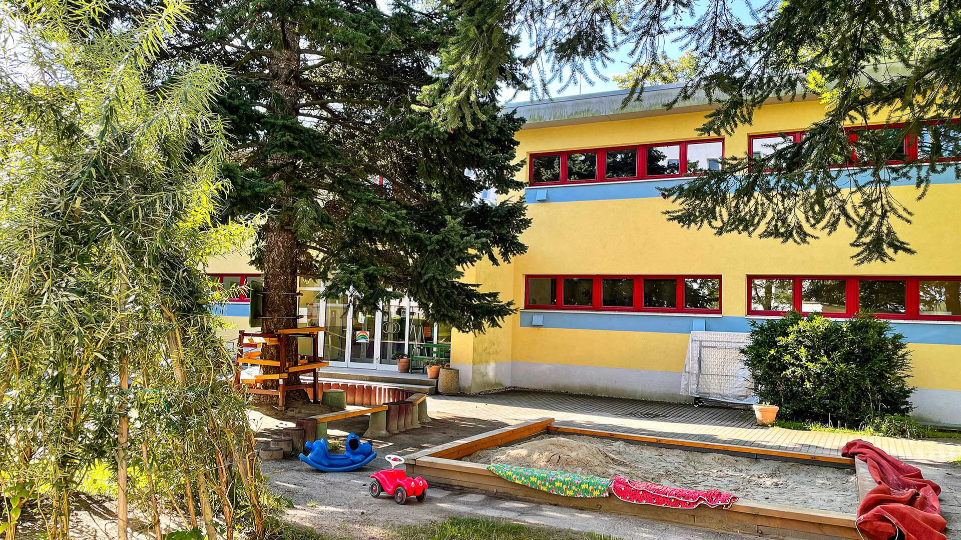 Das Außengelände im Eingangsbereich mit einem Sandkasten, einem Tipi aus Weide und einem Sitzbereich unter einem Baum.