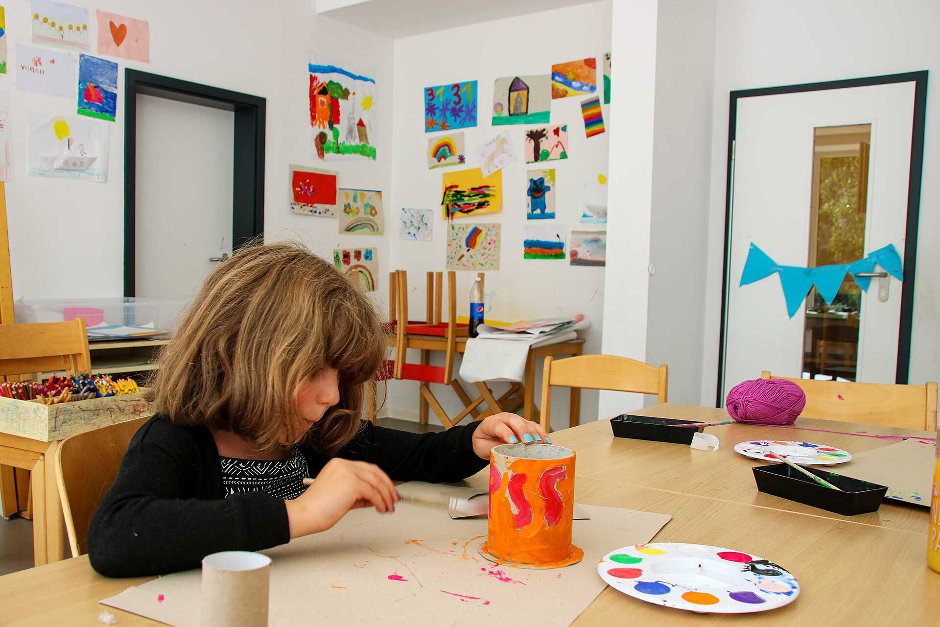 Im Kreativzimmer sitzt ein Mädchen und bemalt eine Dose. Im Hintergrund an der Wand sind viele Kunstwerke zu sehen.