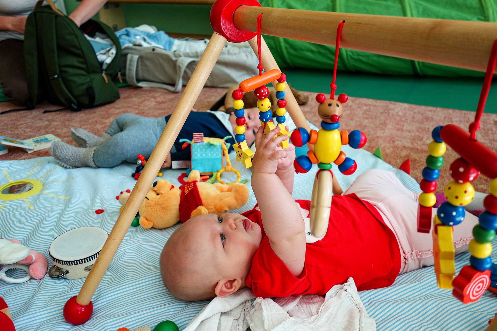 Krabbelgruppe Zwei Babys liegen rücklinks auf einer Decke auf dem Fußboden und greifen nach verschiedenen Spielzeugen.